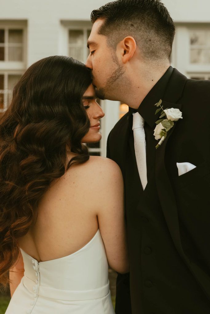 Groom kisses bride's forehead outdoors, bride styled in vintage-inspired waves and natural glam makeup for her wedding day.