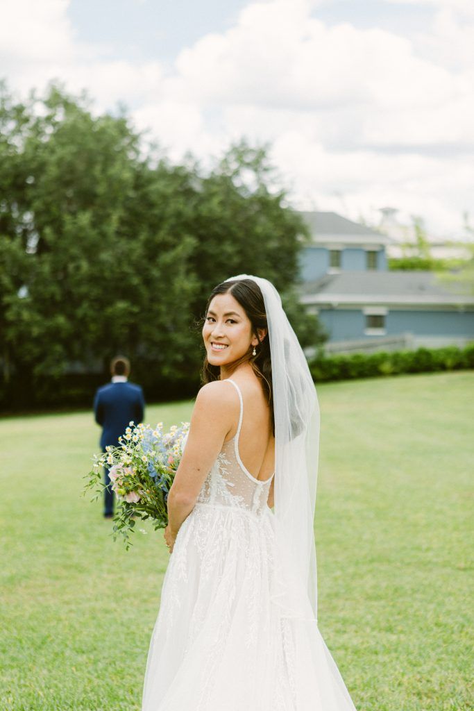 Bride in lace gown and veil holding bouquet, smiling before first look moment, styled with glowing skin and soft waves.