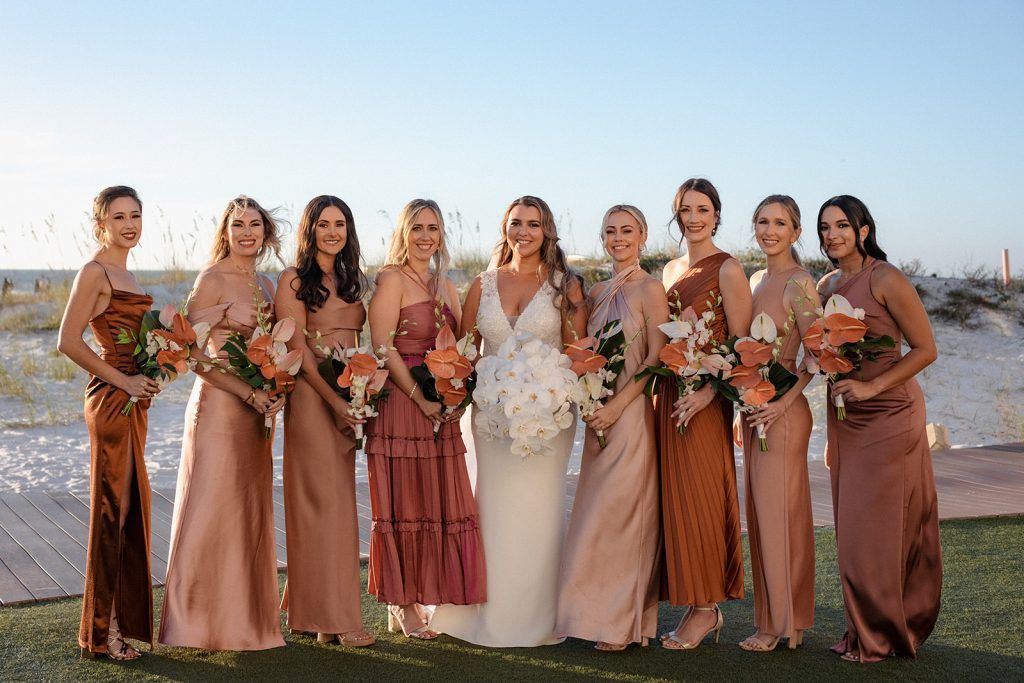 The bride stands in the middle of her bridesmaids on the beach, each holding orange and white tropical bouquets, all smiling at the camera.