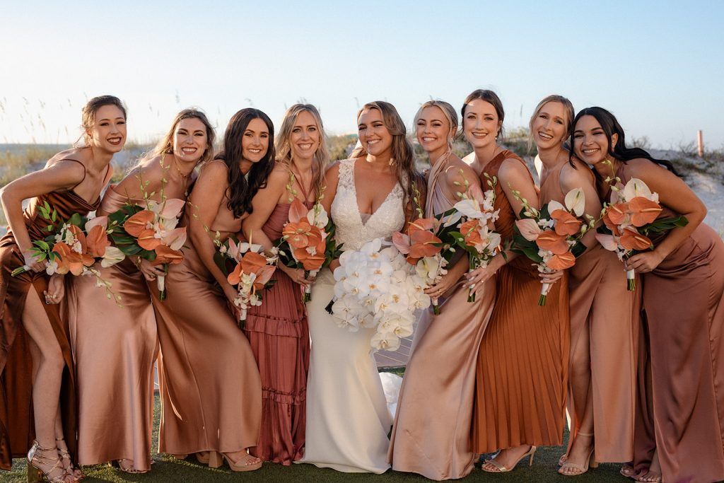 The bride surrounded by her bridesmaids leans in for a playful, joyful group photo, all holding bright orange tropical bouquets.