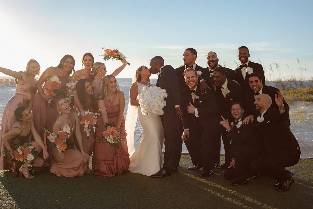 The bride and groom kiss at the center while bridesmaids in rose gowns and groomsmen in black tuxedos celebrate around them on the beach.
