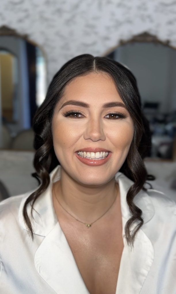 Close-up of a woman with curled hair and natural glam makeup, smiling brightly in a white robe.
