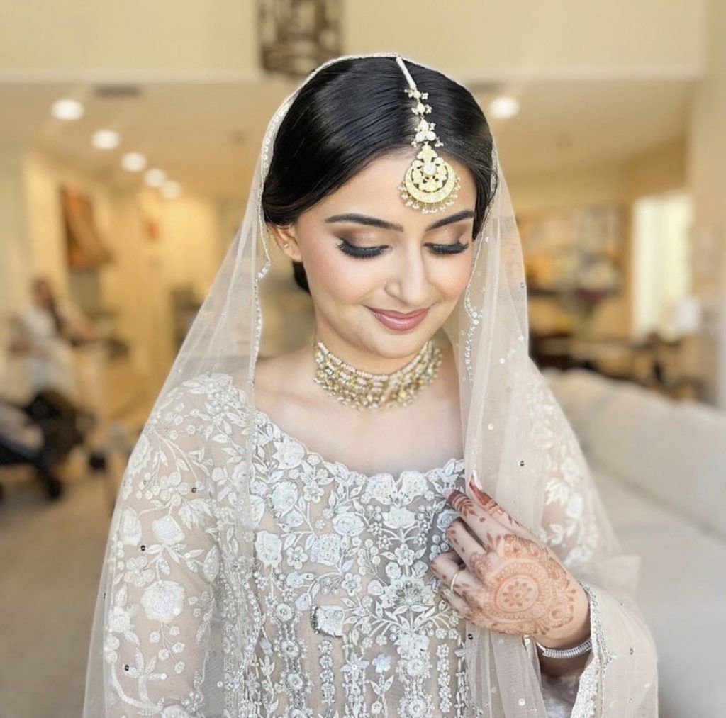 Bride in an embroidered ivory outfit with a jeweled headpiece, veil, and intricate henna on her hand, smiling softly while looking down.