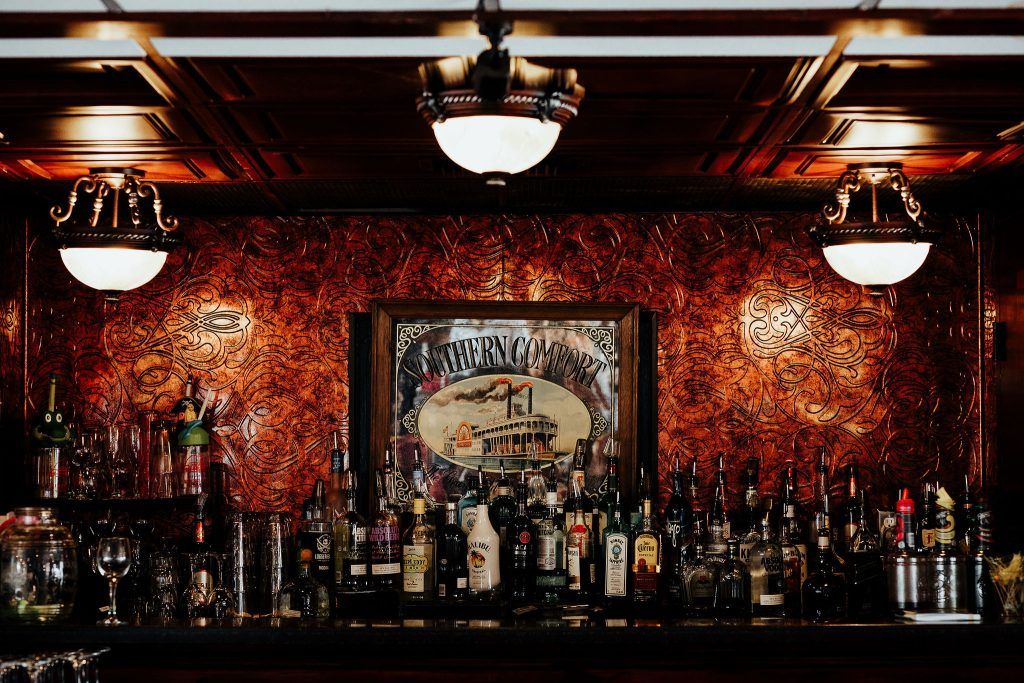 A bar with a decorative red patterned wall, liquor bottles lined up, and a framed "southern comfort" steamboat painting in the center.