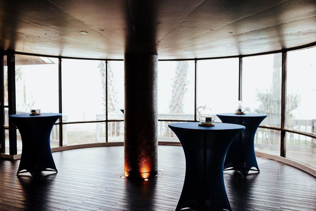 Cocktail tables with navy-blue covers and glass centerpieces arranged in a room with floor-to-ceiling windows and waterfront views.
