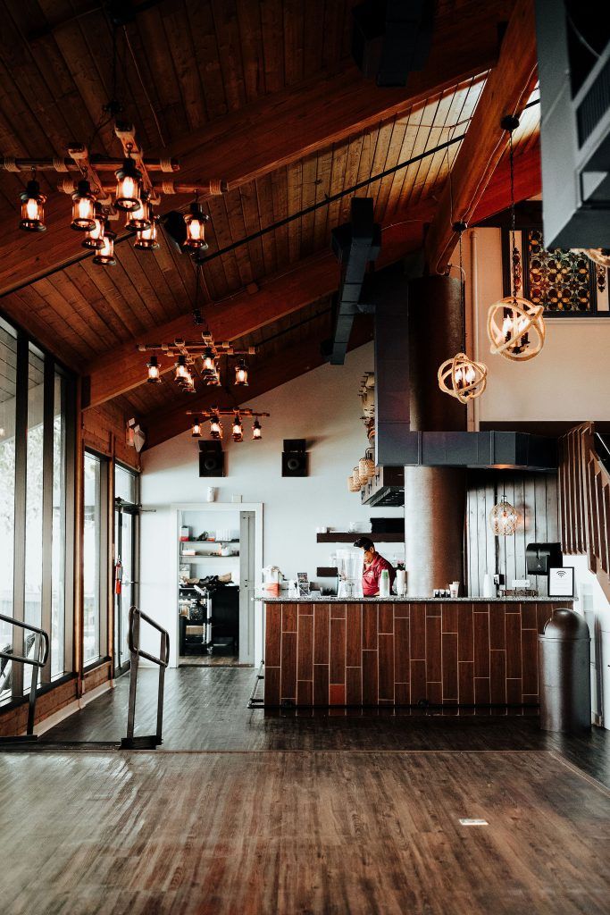 Warm wood interior with rustic chandeliers, a bar counter with a staff member behind it, and large windows letting in natural light.