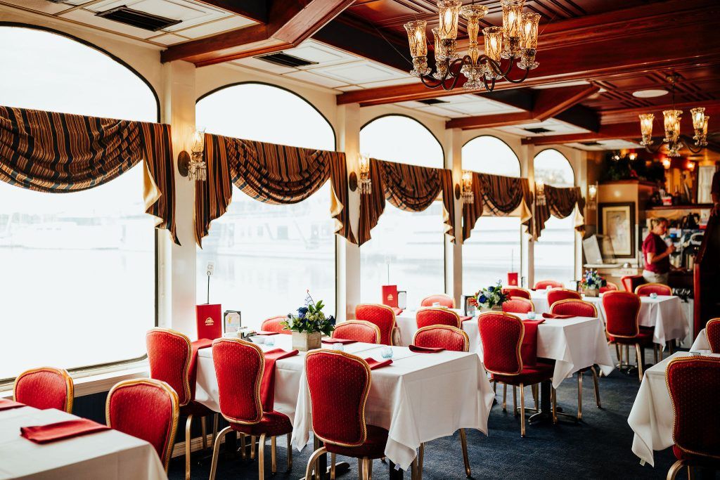 Indoor dining area with a waterfront view, featuring a table with red chairs, white tablecloth, and nautical-themed decor.