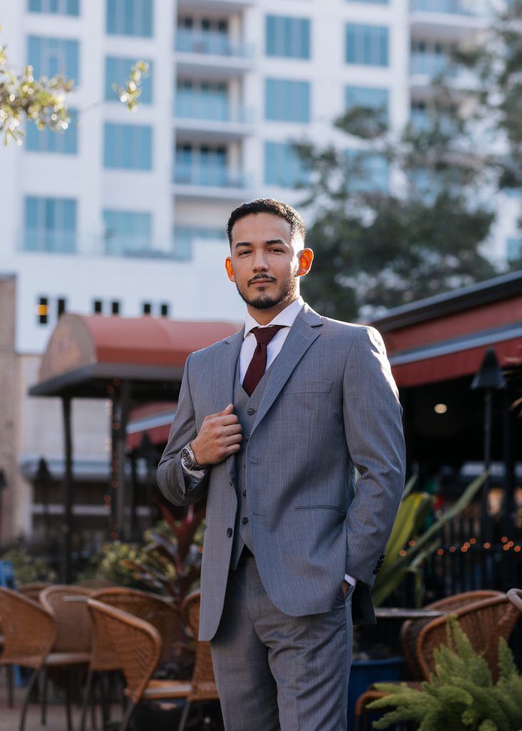 Man wearing a light grey suit with a brown tie looks at the camera