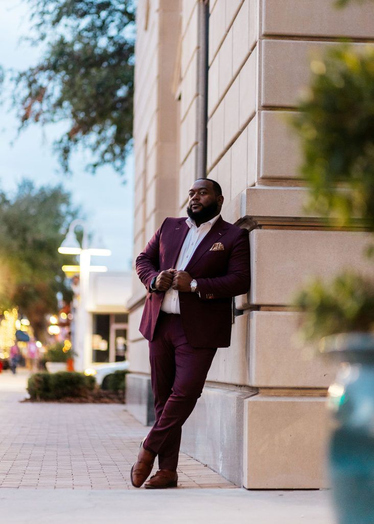 a man wearing a dark maroon suit leans against a tan wall