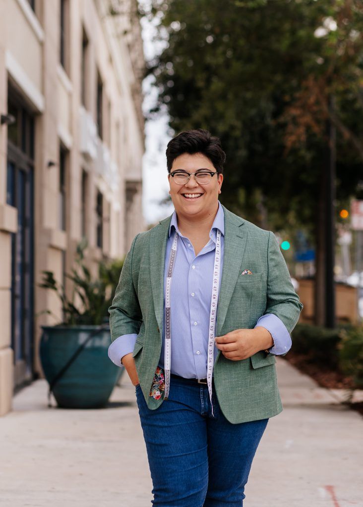 a suit tailor smiles at the camera with a tape measure around their neck