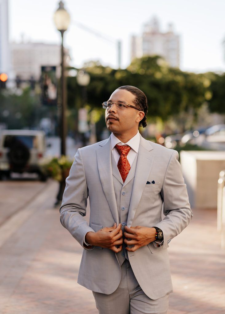man looks off into the distance to his right wearing a light grey suit with a patterned red tie
