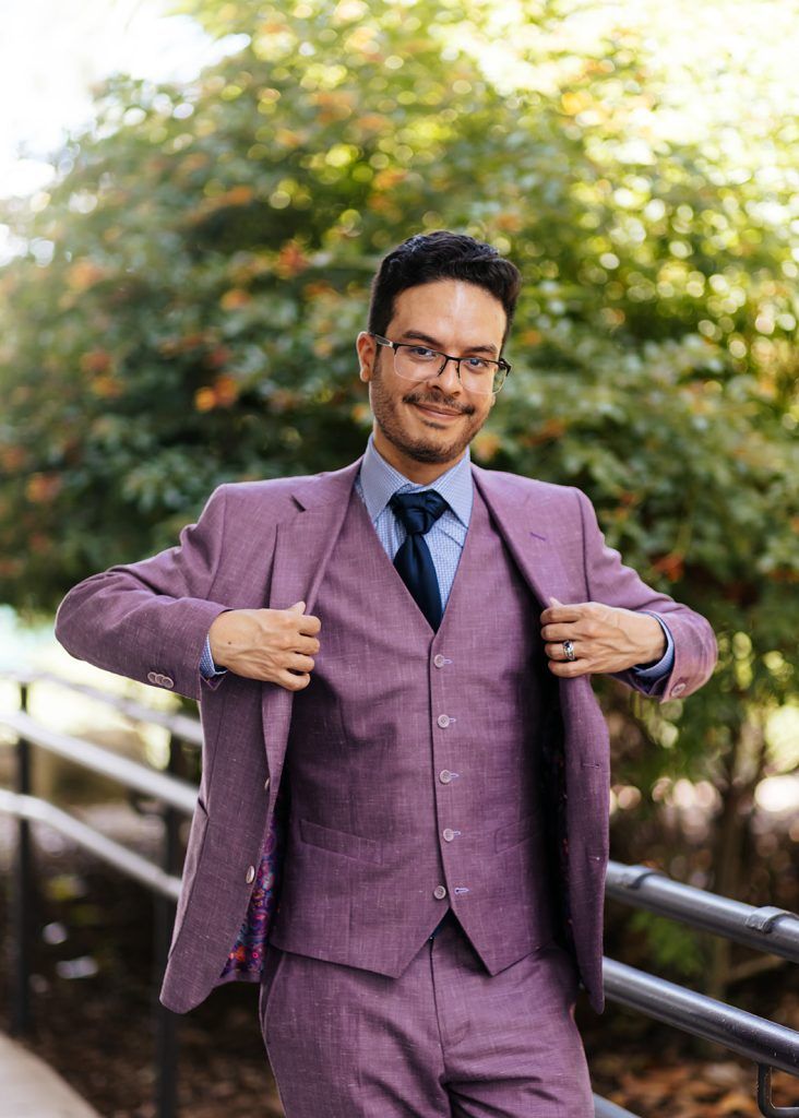 man wearing a plum suit with a blue undershirt and black tie smiles at the camera