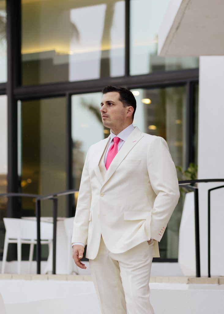man looks off into the distance wearing an off white suit, white undershirt, and pink tie