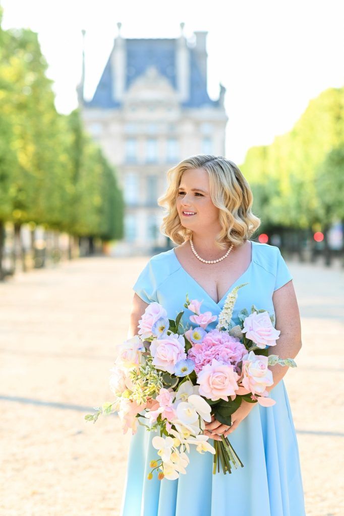 A smiling bride in a light blue dress holds a large pastel bouquet