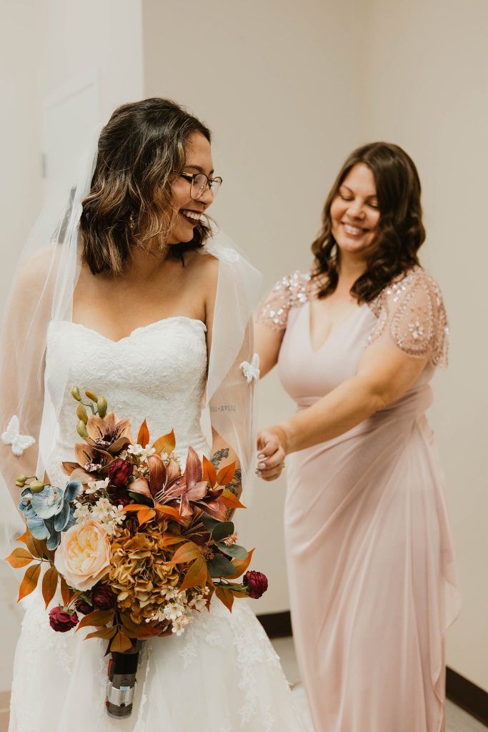 a bride smiling as another woman adjusts her dress, holding a fall-inspired bouquet