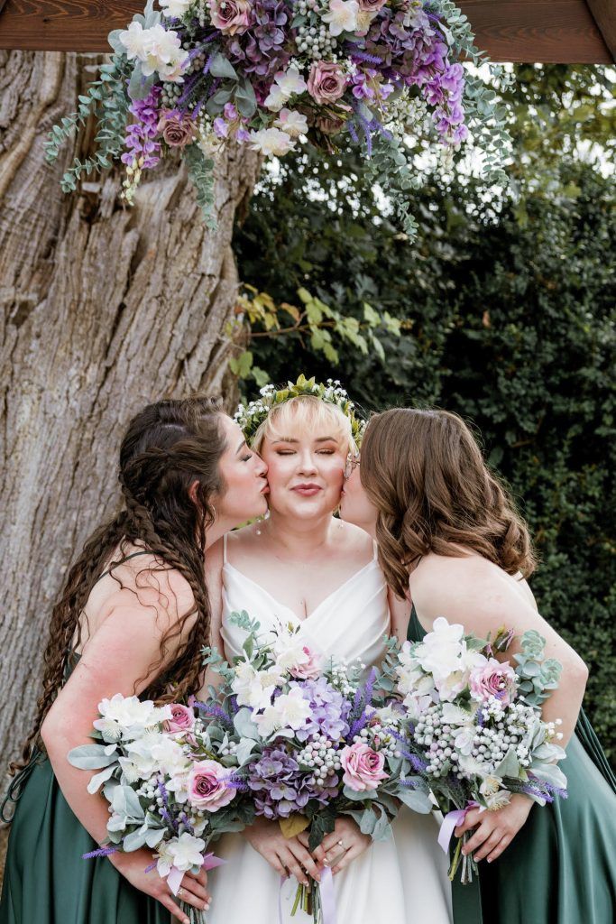 a bride standing between two bridesmaids, each holding purple, pink, & white bouquets